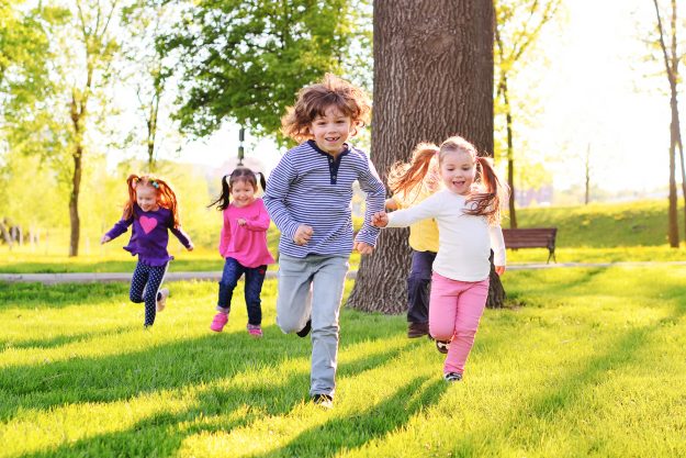 a group of small happy children run through the park in the background of grass and trees.